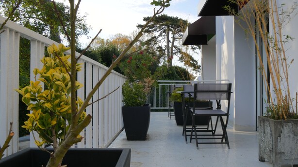 An outdoor patio with a white fence, potted plants, a table with chairs, and a building with a dark facade. - Hôtel Savary (La Rochelle)