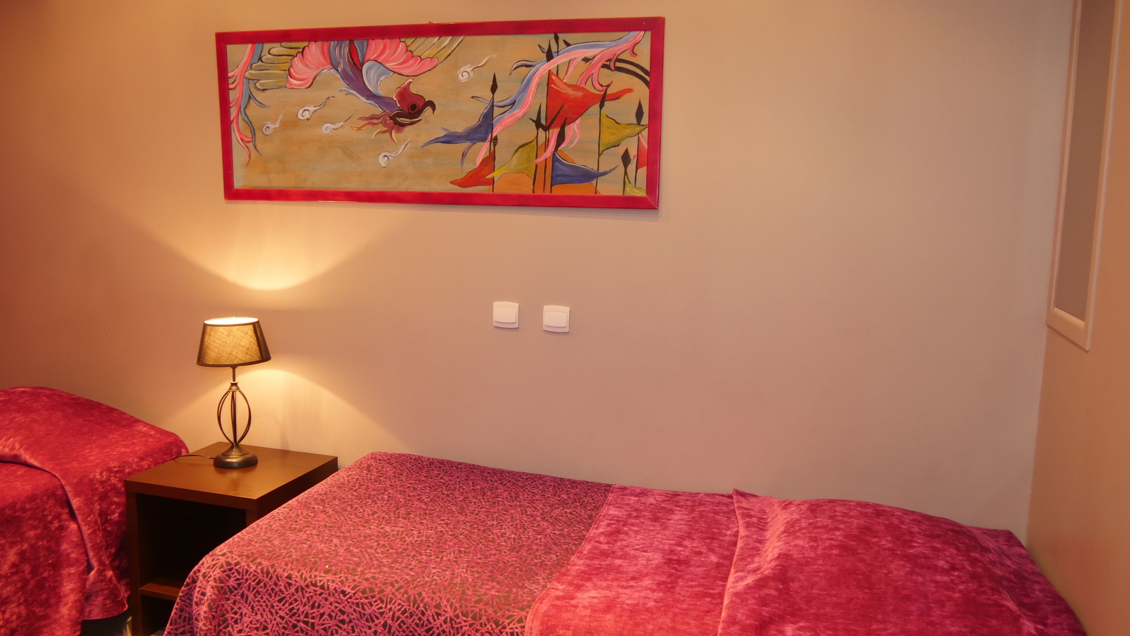 A room with two beds, a nightstand with a lamp, a wall-mounted painting, and two wall sockets.
