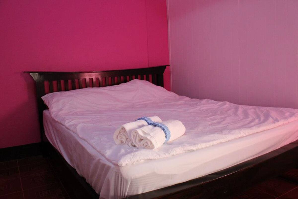 A neatly made bed with white linens and a rolled towel on top, set against a pink wall.