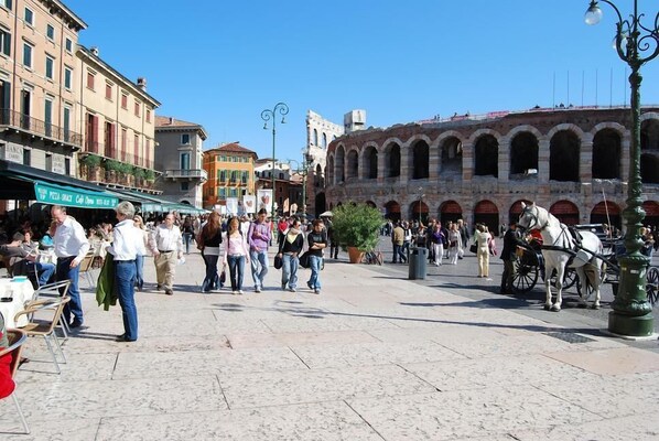 A bustling square with historical architecture, including a prominent archway and a horse-drawn carriage. - Art&Coffee (Verona)