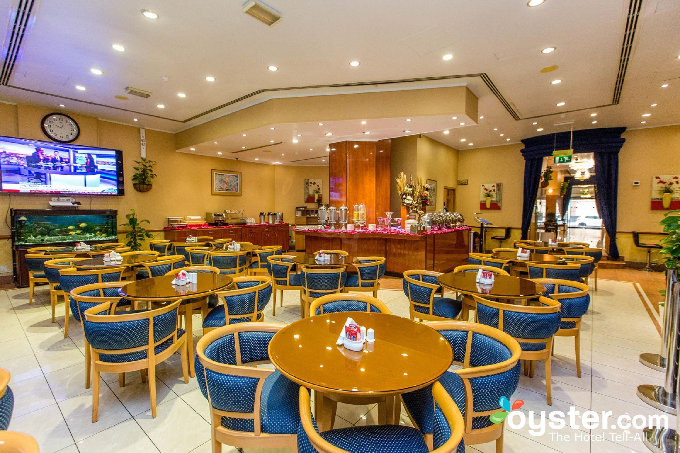 A hotel lobby with round tables and blue chairs, a reception desk, and a television displaying news.