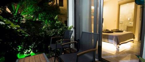 Un balcon avec une table et des chaises, un lit visible à travers une porte vitrée et un espace jardin illuminé de lumière verte.