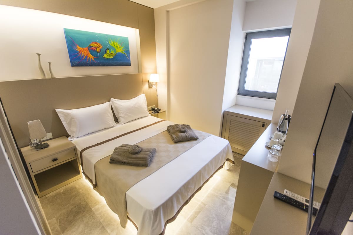 A hotel room with a bed, bedside table, lamp, artwork, window, and a TV.
