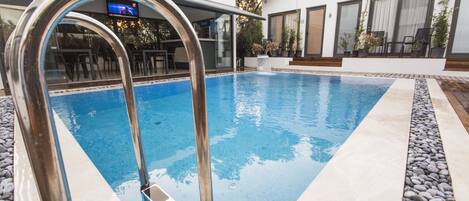 A swimming pool with a metal ladder, surrounded by a stone border, and adjacent to a modern building with large windows and outdoor seating.