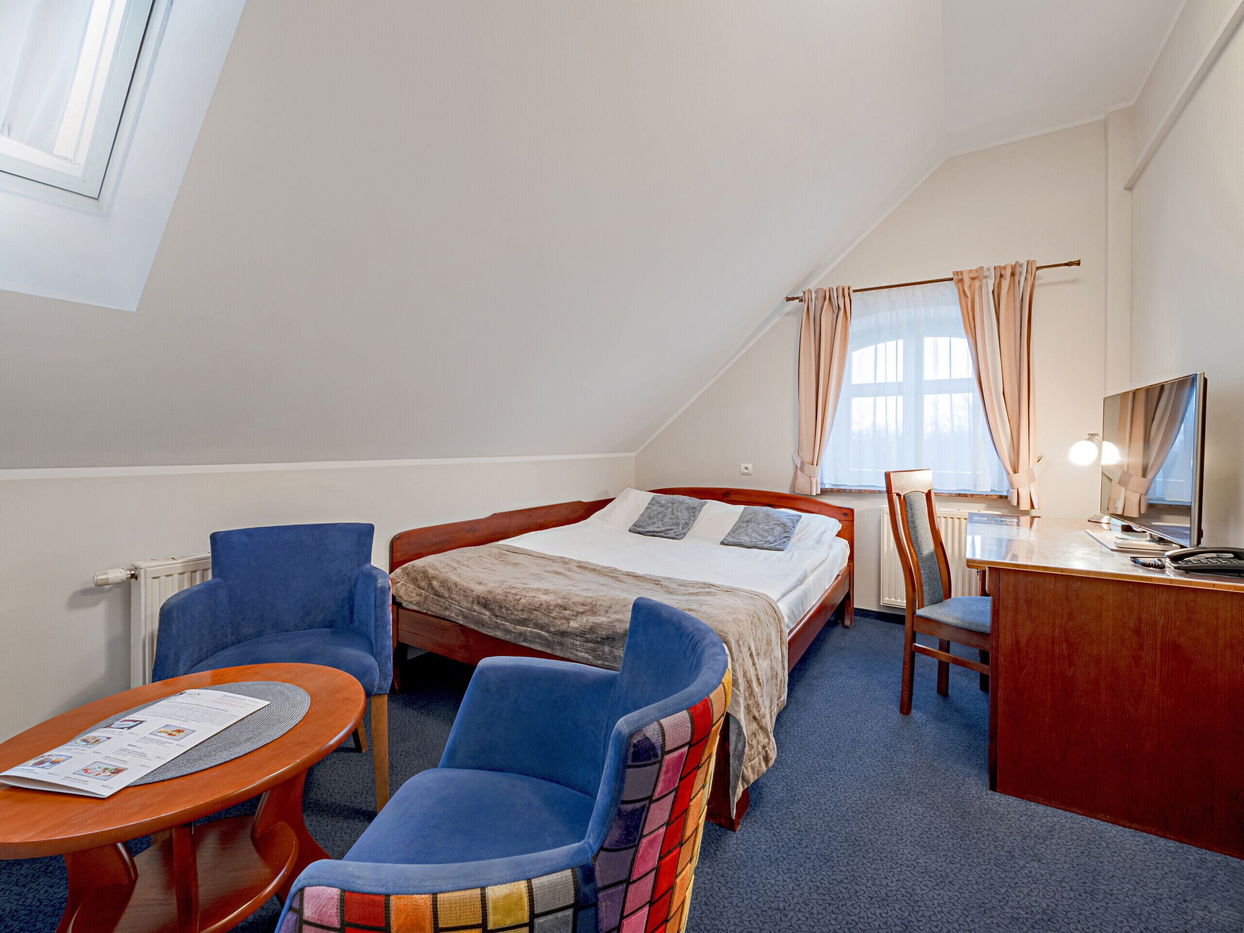 A hotel room with a bed, two blue chairs, a small wooden table, a desk with a chair, a TV, and a window with curtains.