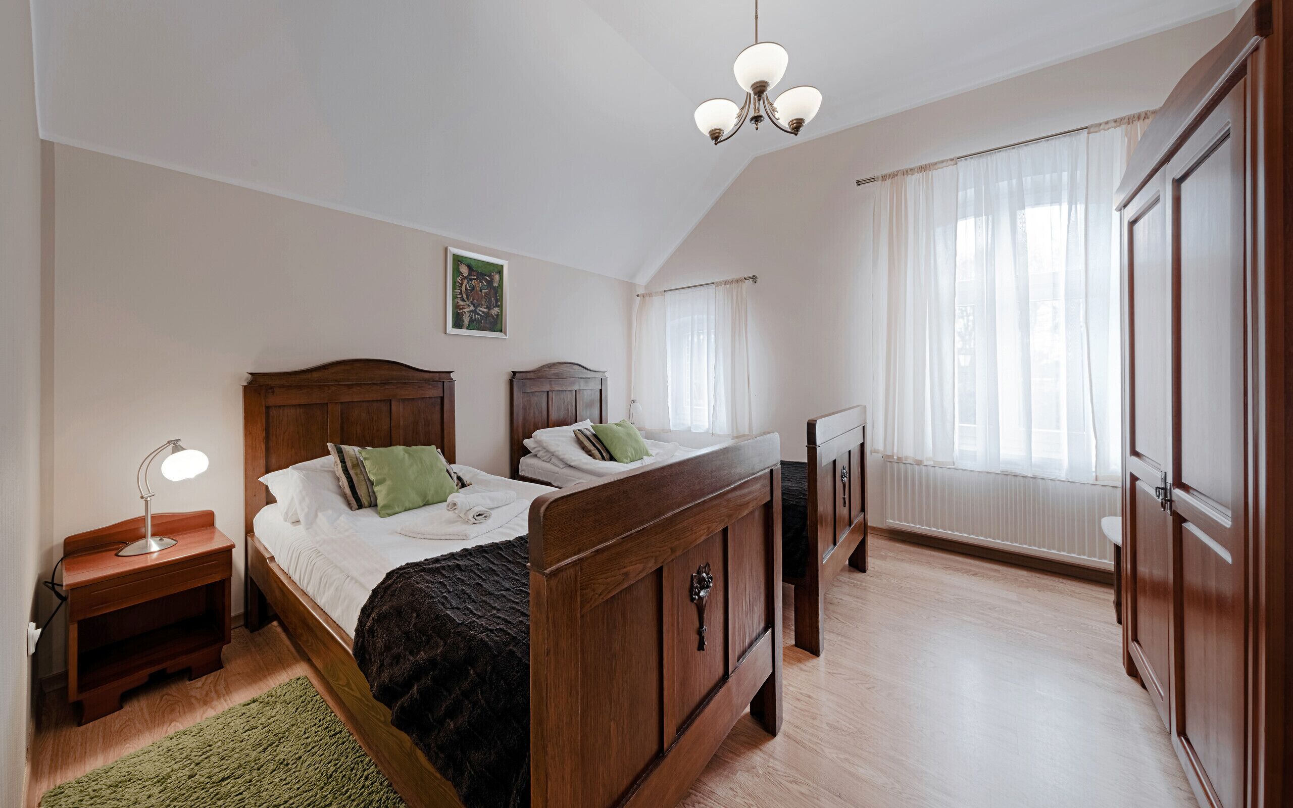 A bedroom with a wooden bed, a nightstand, a desk, a radiator, and a window with curtains.