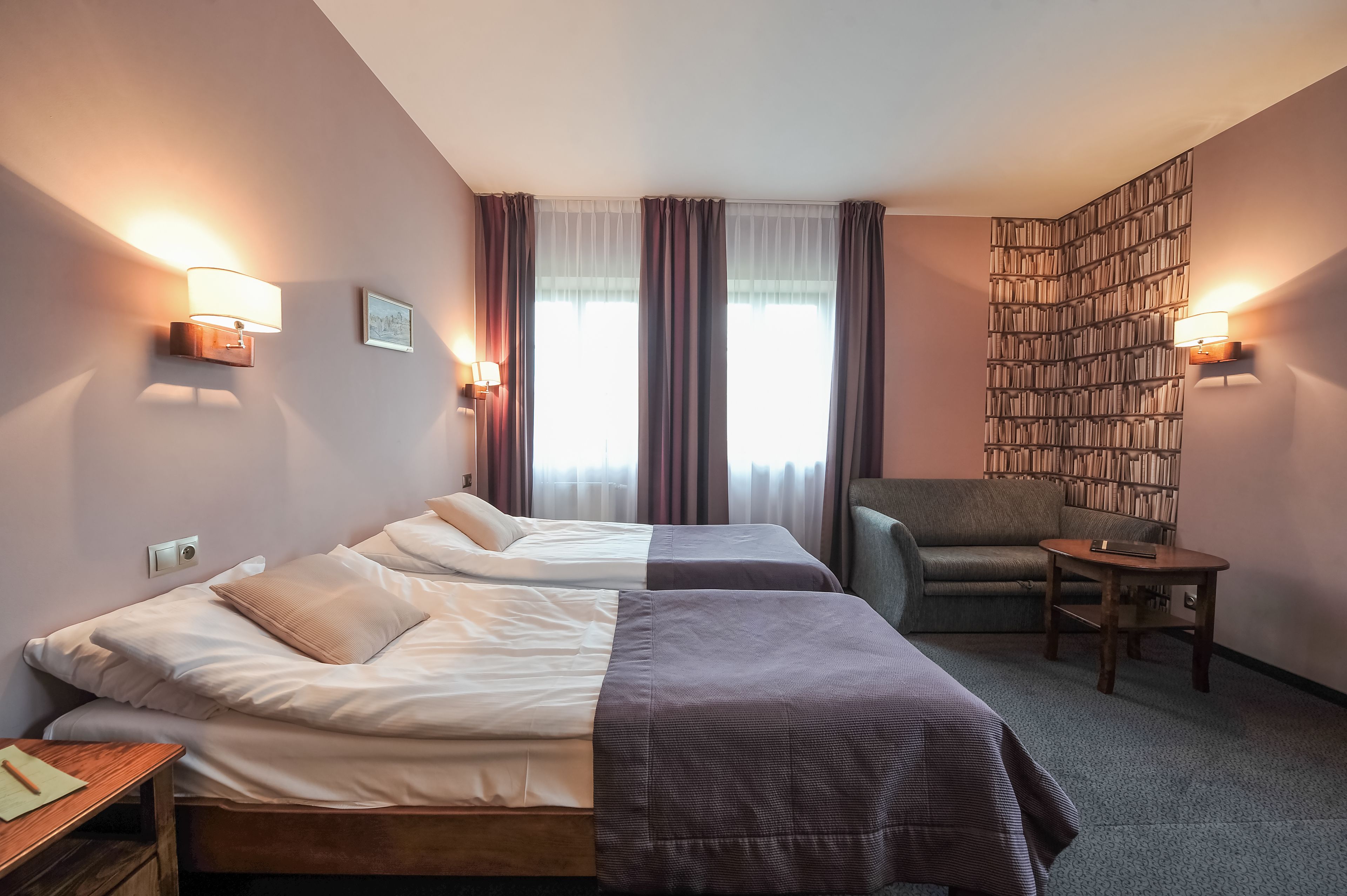 A hotel room with two beds, a bookshelf, a chair, and a small table.