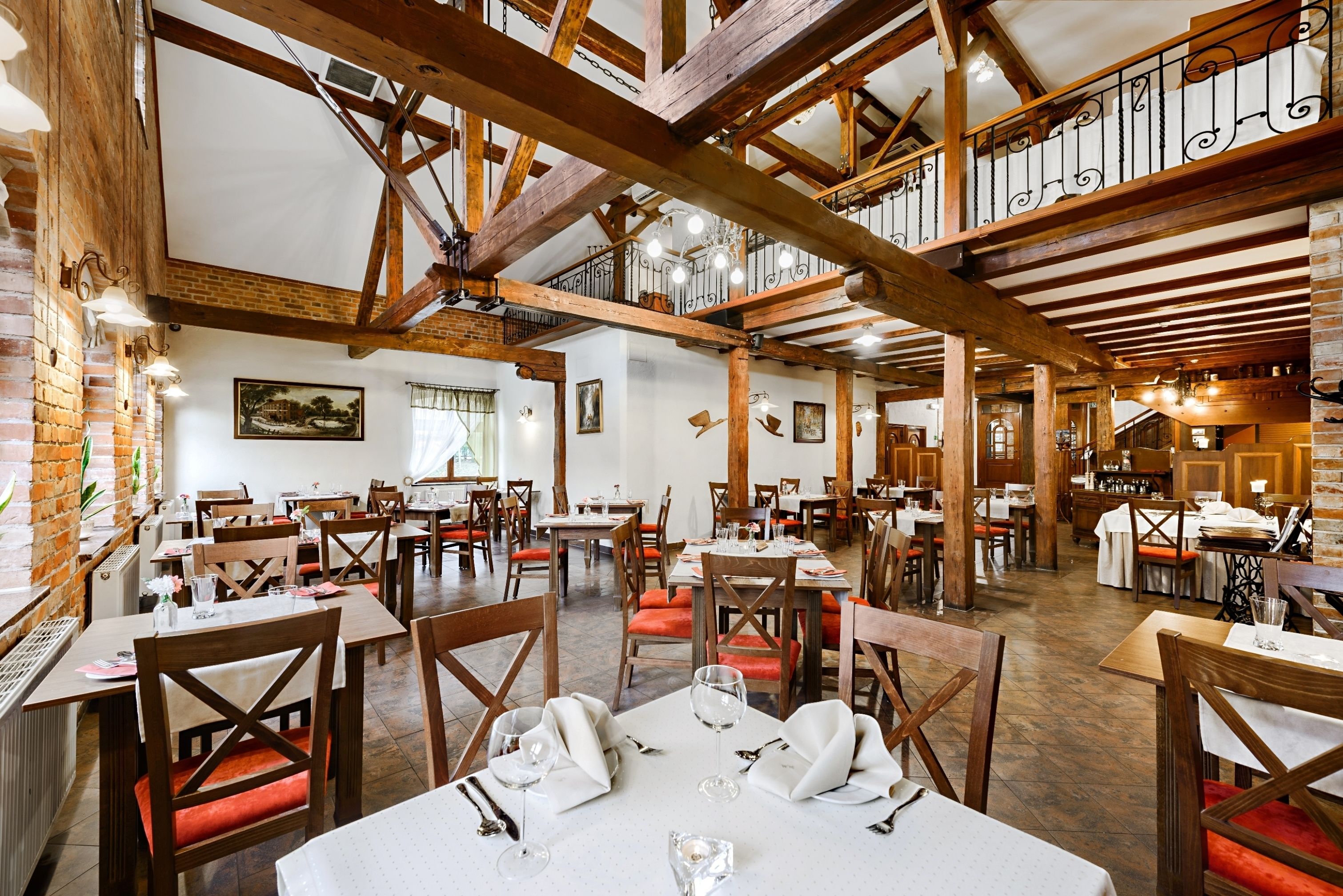 A spacious restaurant with high wooden beams, multiple dining tables, and red cushioned chairs.