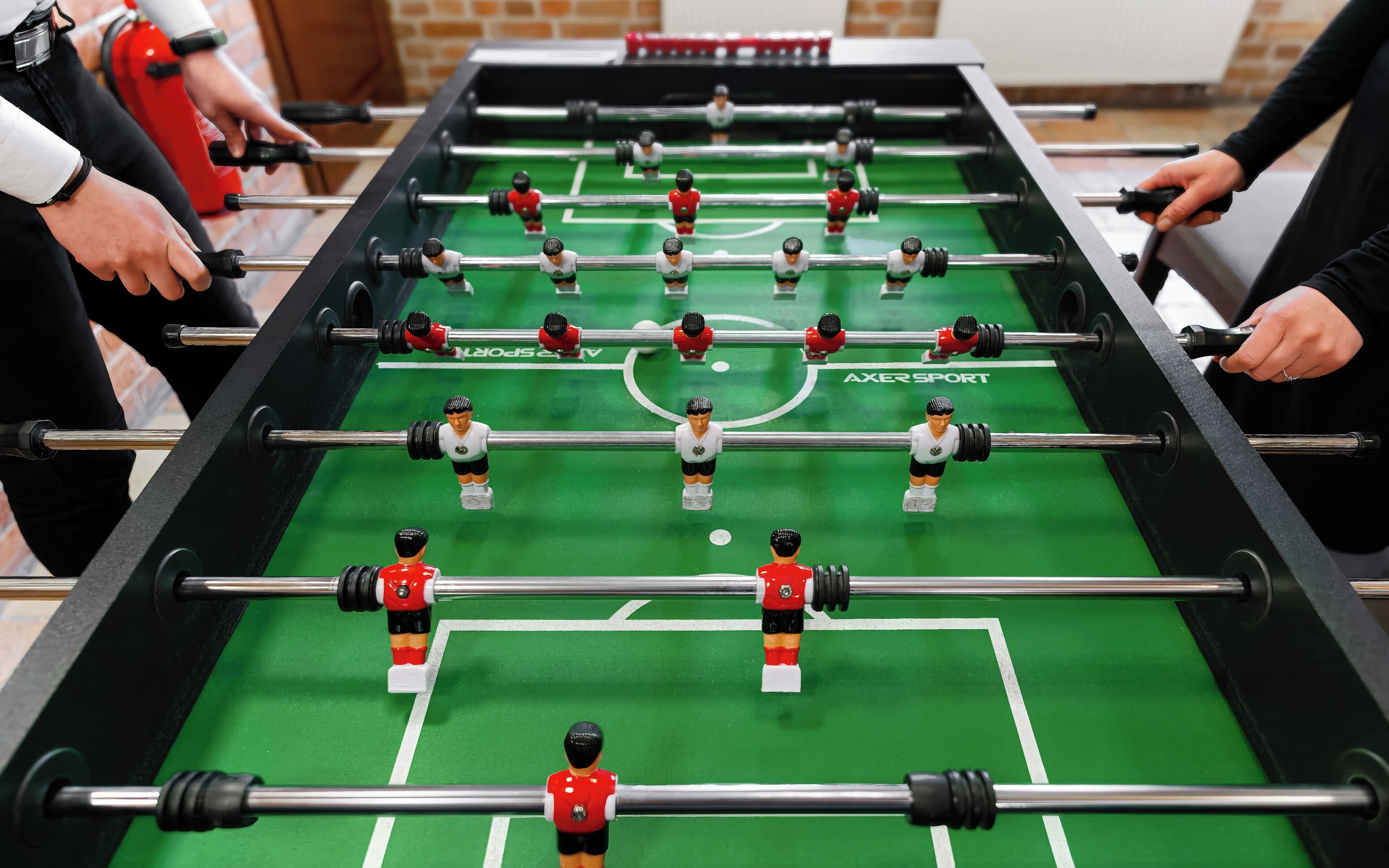Ein Kicker mit Spielfiguren in roten und weißen Trikots, in einem Raum mit Backsteinwänden, im Hintergrund ist ein Feuerlöscher zu sehen.