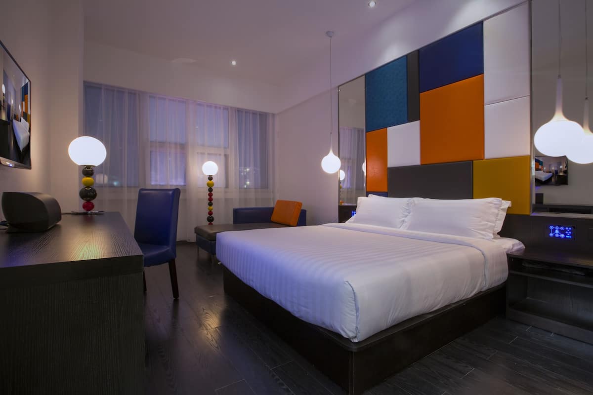 A modern hotel room with a large bed, a desk, and a chair. There is a mirror on the wall and a window with curtains.