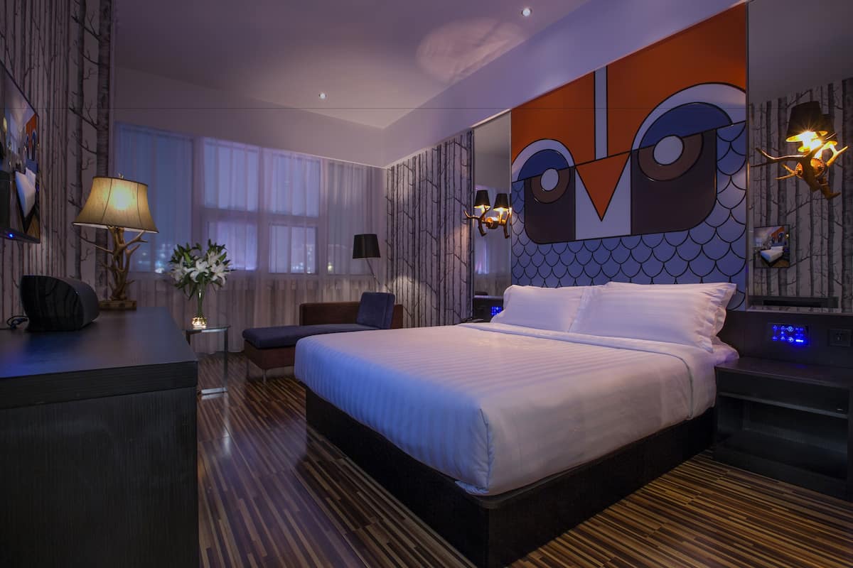 A modern hotel room with a large bed, a desk, a chair, and a nightstand with a digital clock.
