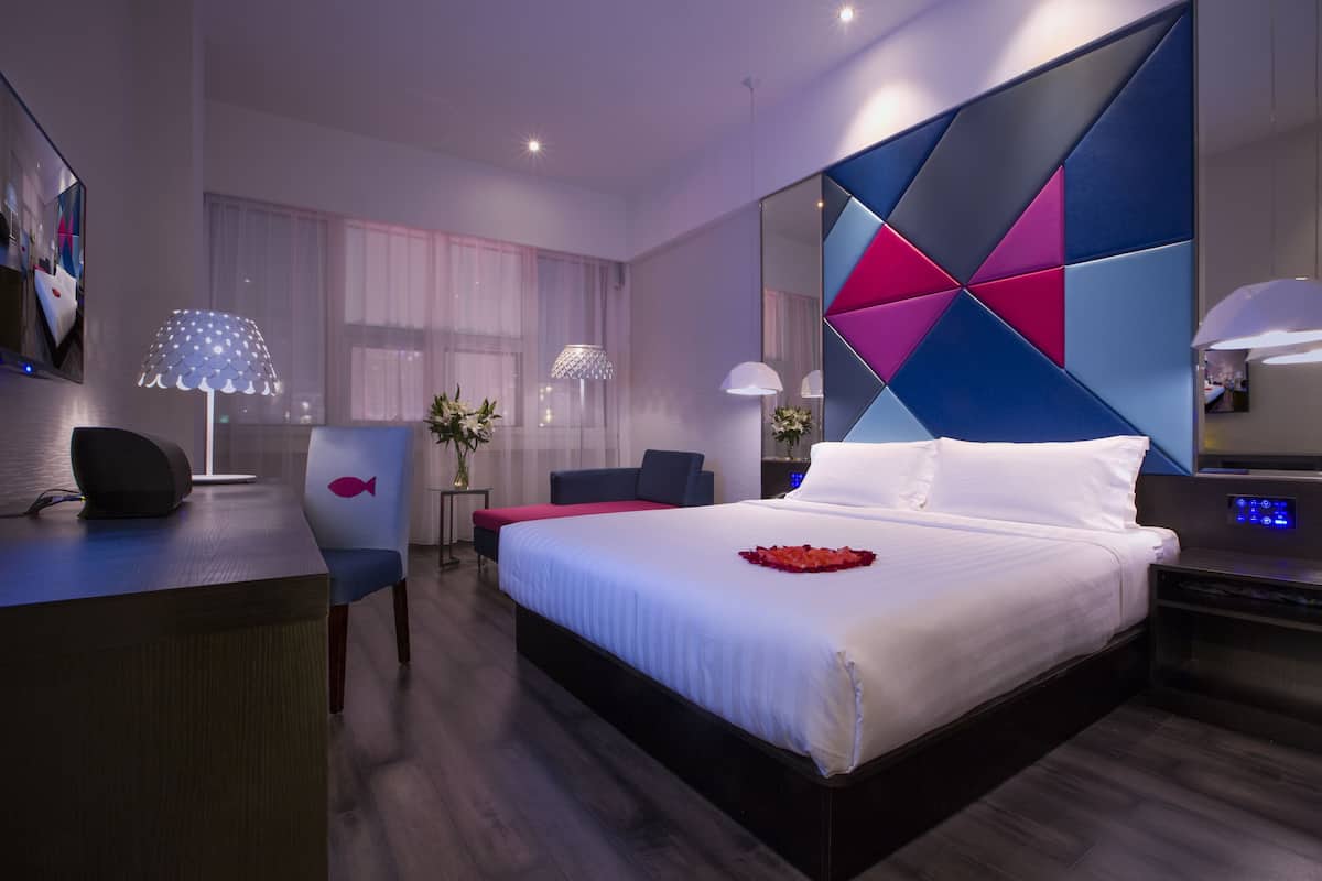 A modern hotel room with a large bed, a desk, a chair, and a bedside table.