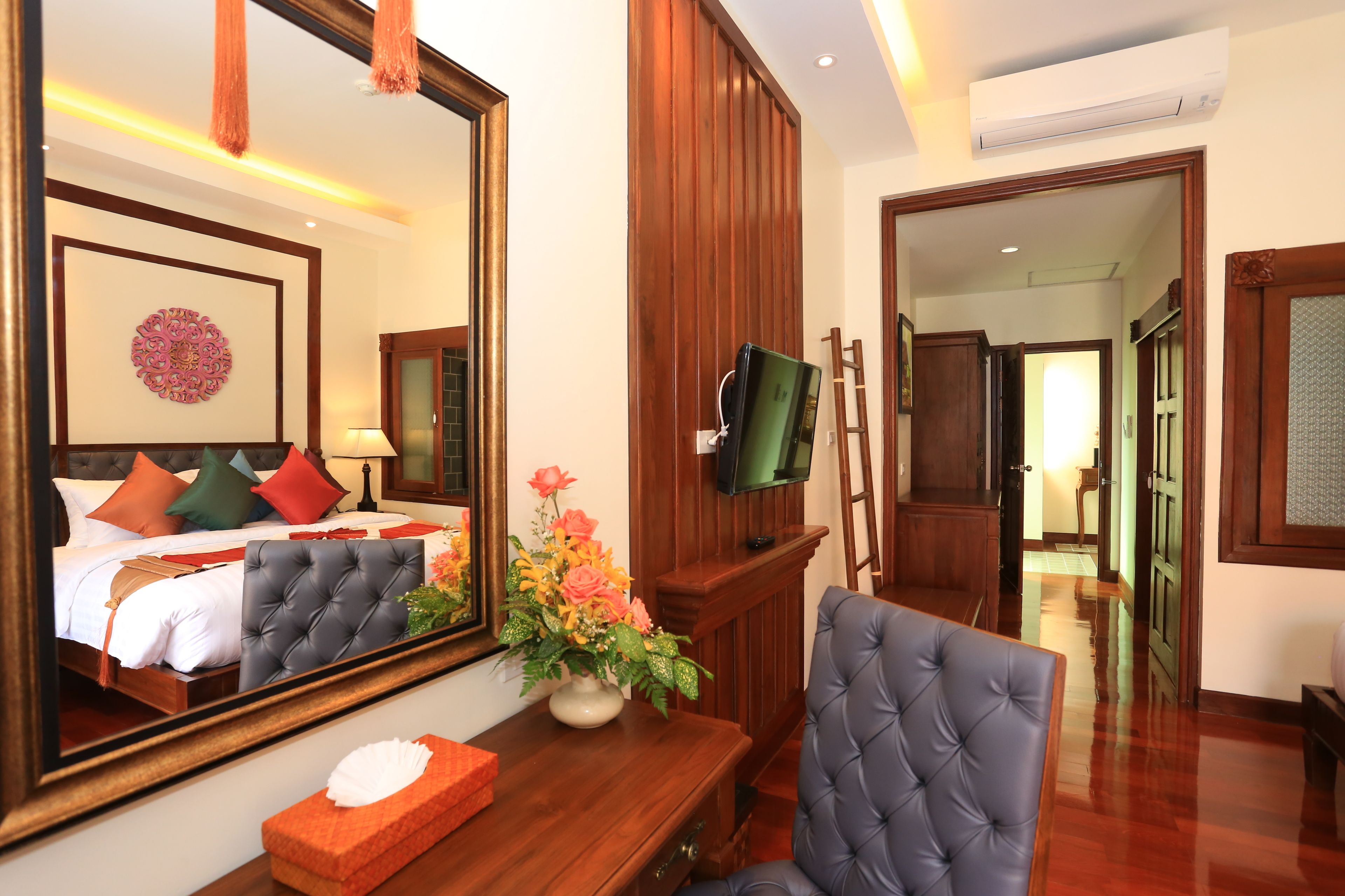 A hotel room with a bed, a large mirror, a television, a wooden desk, a chair, and a vase of flowers.