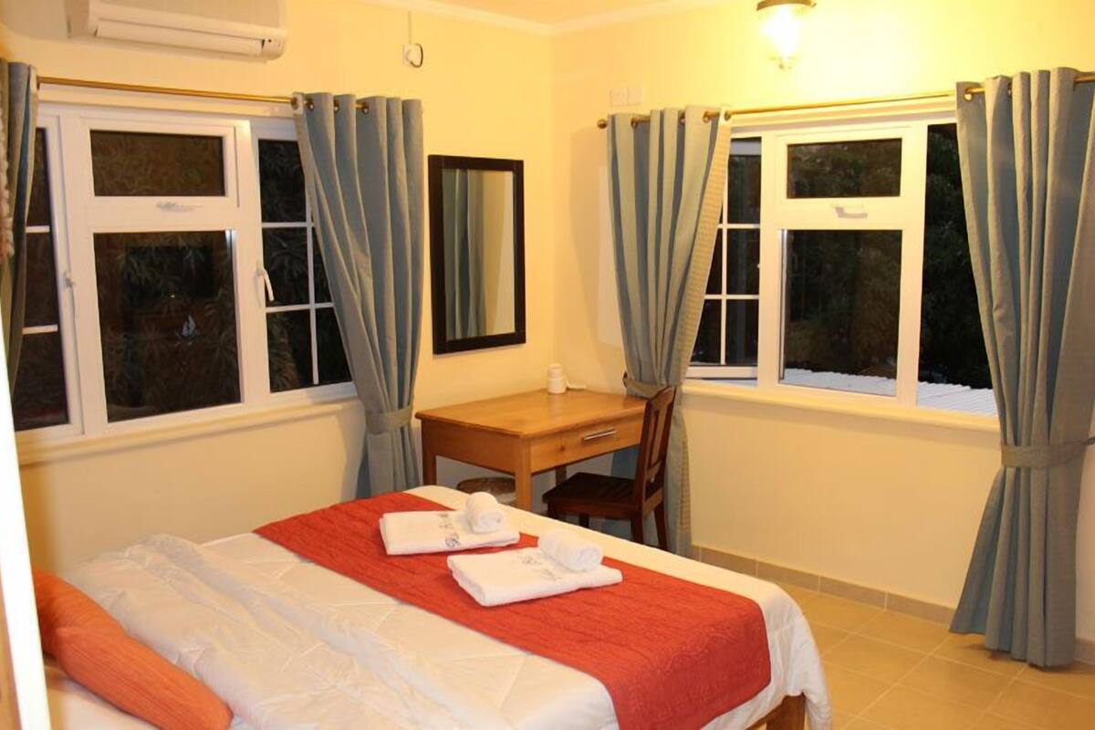 A hotel room with a bed, a desk, and two windows with curtains.