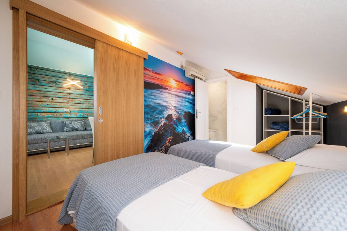 A modern hotel room with two beds, a sofa, a mural of a sunset, and a wooden wardrobe.