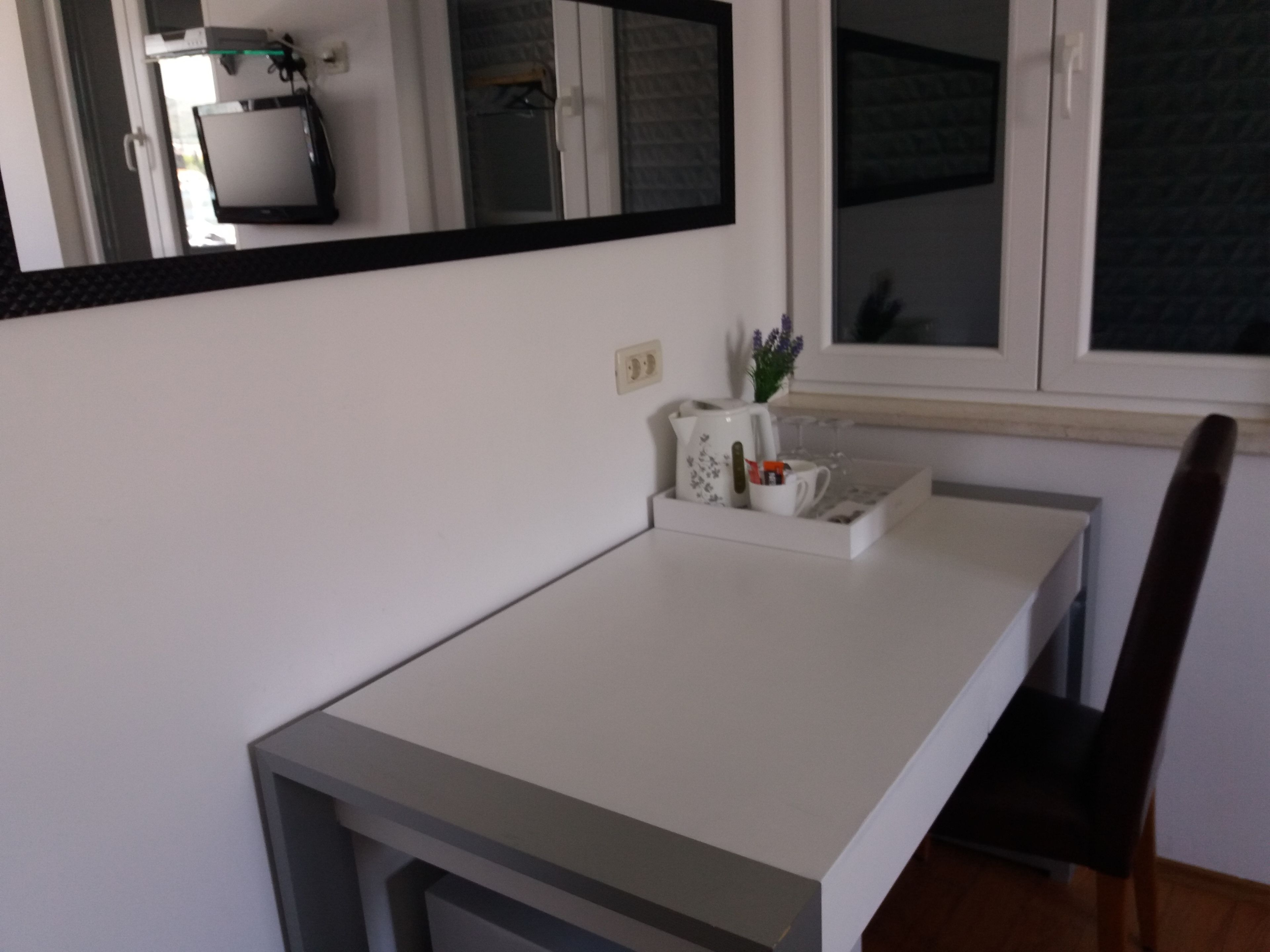 Un escritorio blanco con una bandeja que contiene una taza, un vaso y una pequeña planta. Una silla está ubicada a la derecha del escritorio.