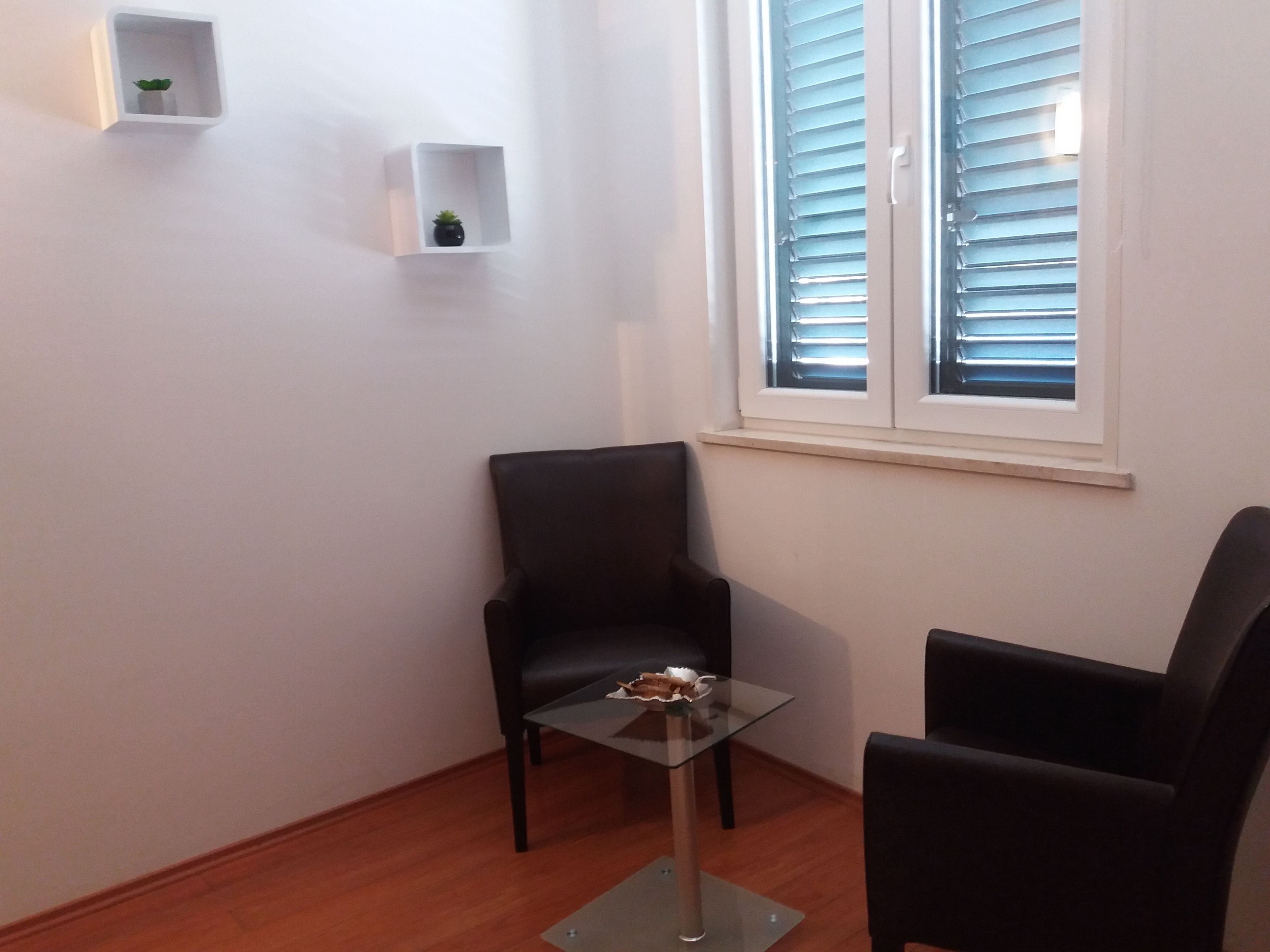 A small room with a glass table, two black chairs, a window with blinds, and two white shelves with plants.