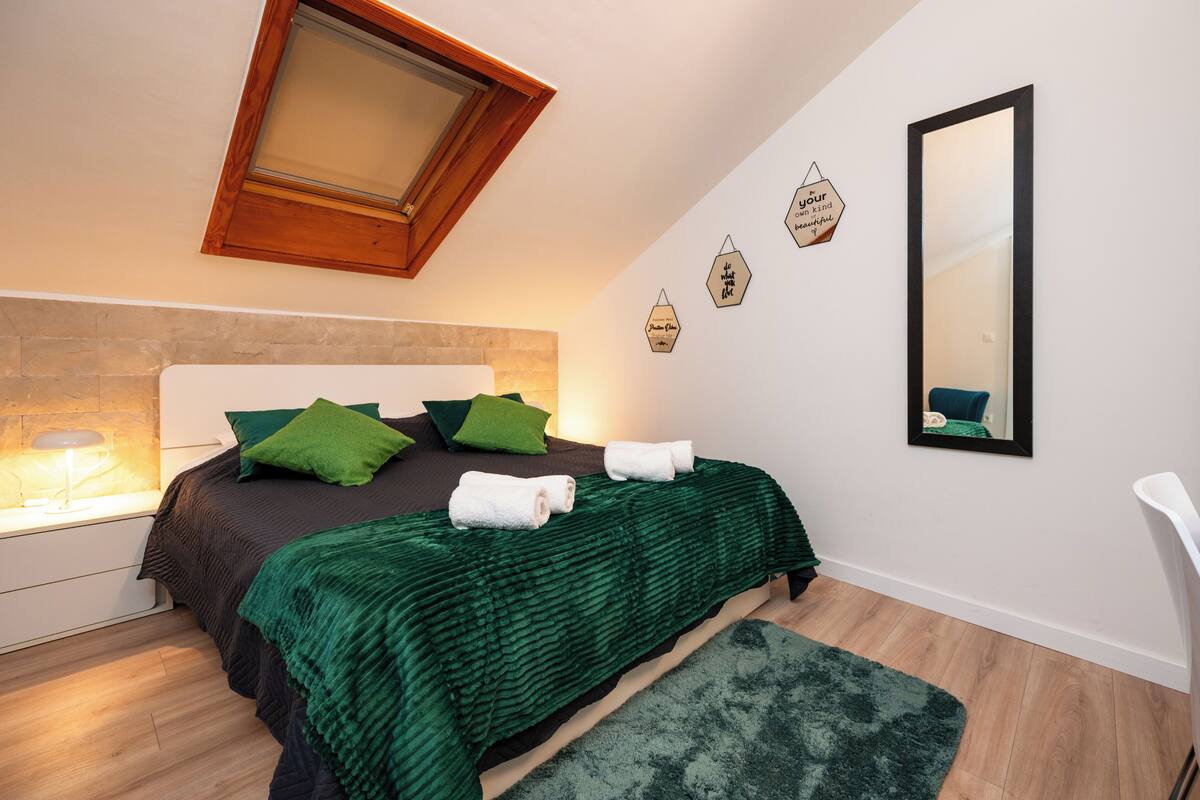 A bedroom with a bed, green pillows, a green blanket, a nightstand with a lamp, a mirror, and hexagonal wall decorations.