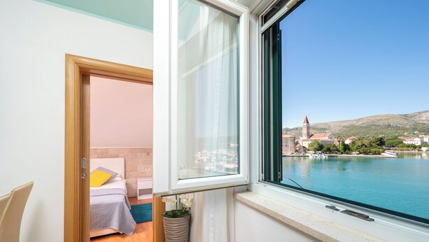 A room with a bed, a window with a view of a body of water and a distant building, and a potted plant.