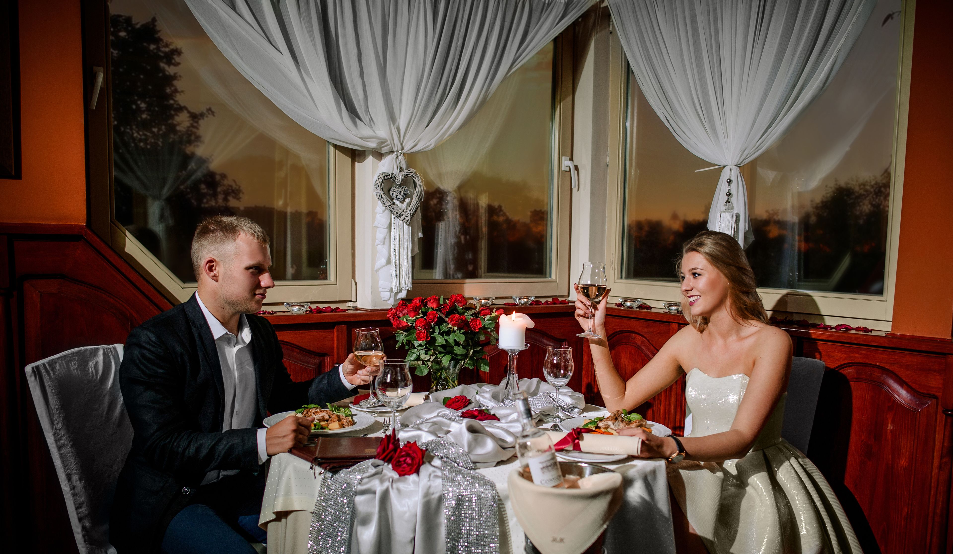 Un couple est assis à une table, devant une bougie, des verres à vin et un bouquet de roses rouges.