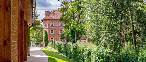 A pathway leading to a brick building with a red roof, surrounded by lush greenery and a green fence.
