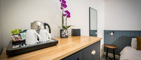 A hotel room with a wooden counter, a coffee machine, a tray with coffee cups and packets, a vase with purple flowers, a bed, and a mirror.