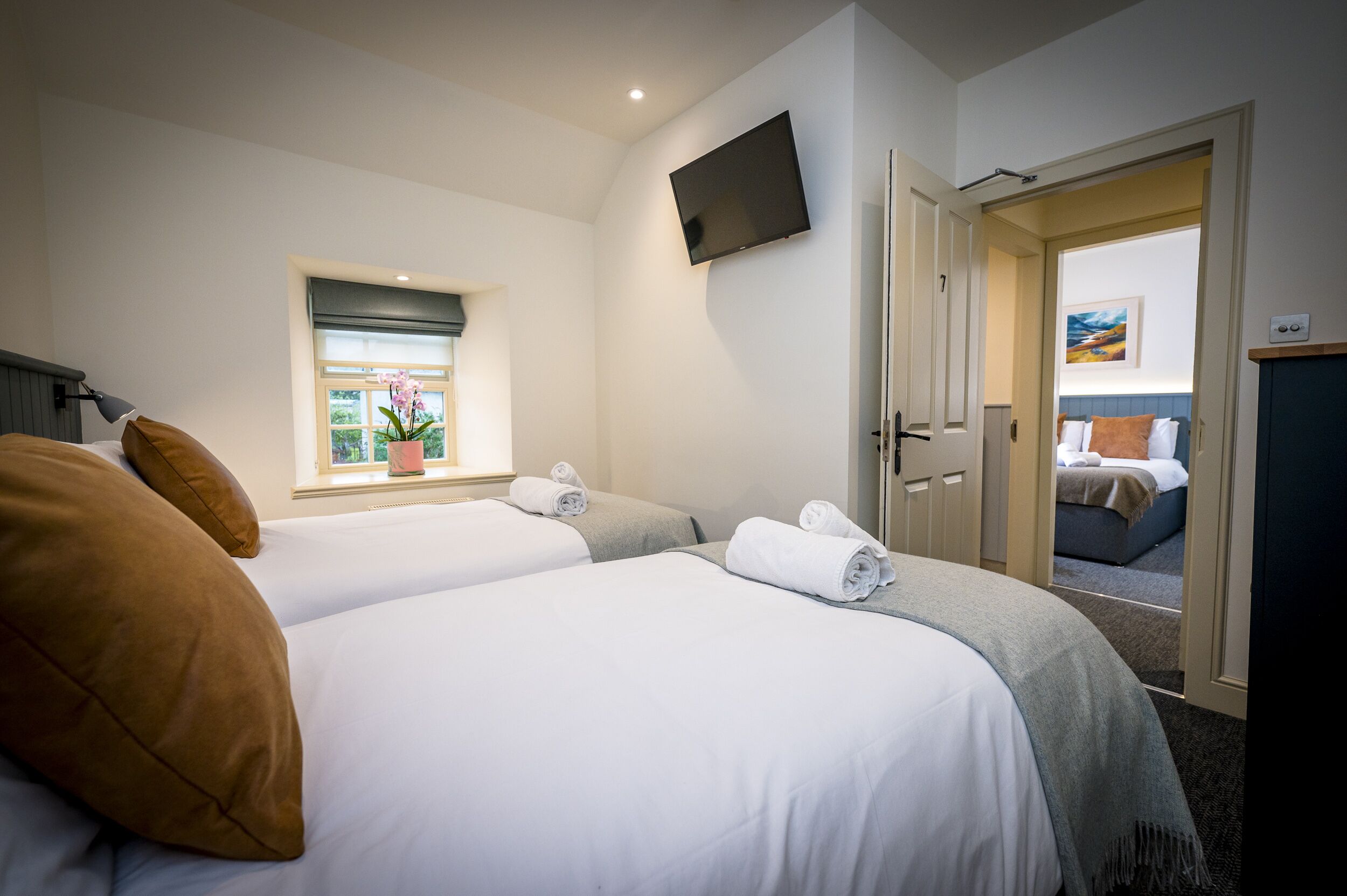 Habitación de hotel con dos camas, un televisor montado en la pared, una ventana con vista a vegetación y una puerta que da a otra habitación.