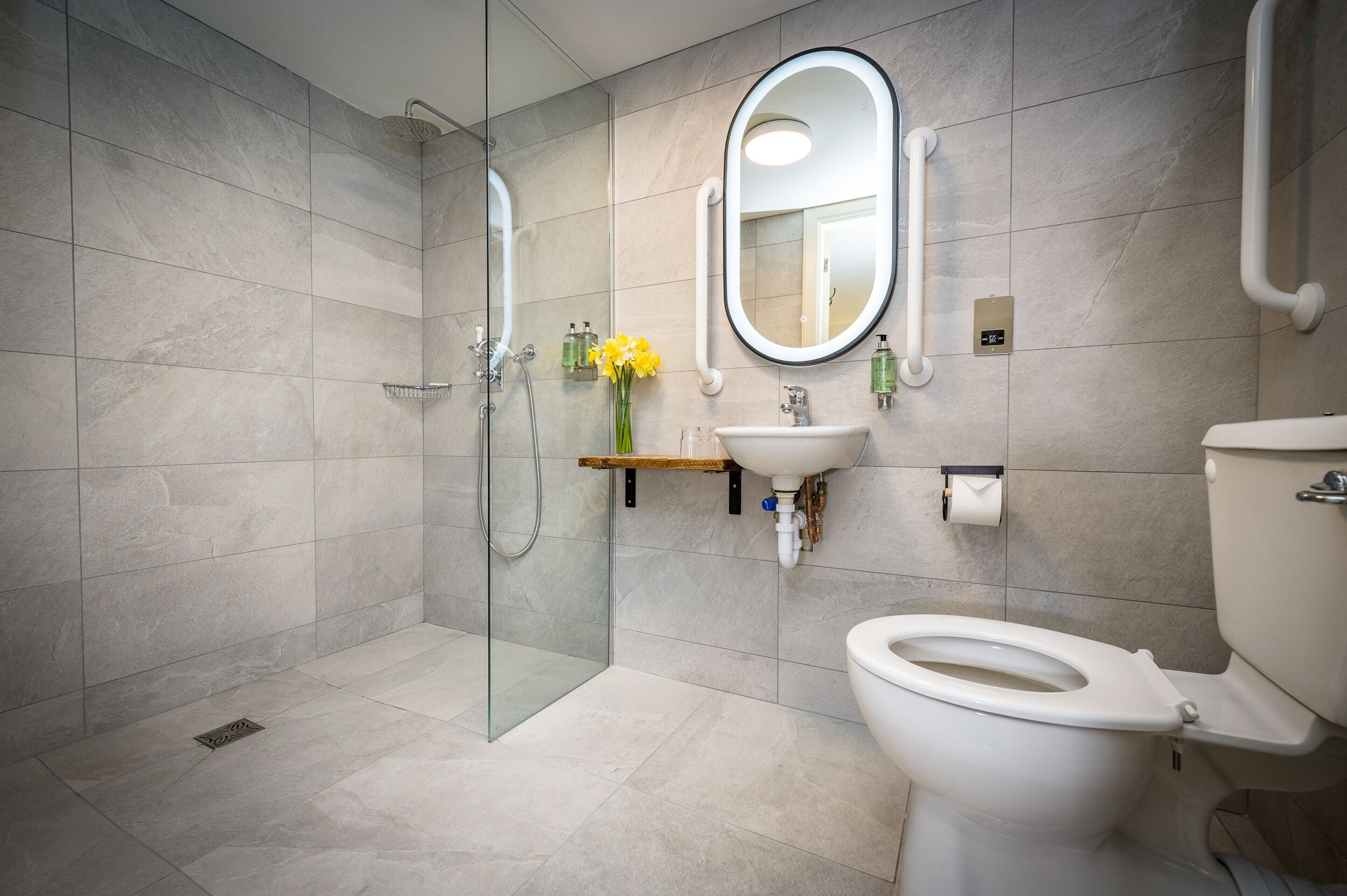 Un baño moderno con ducha envuelta en vidrio, inodoro blanco, lavamanos con espejo y un pequeño estante con artículos de tocador.