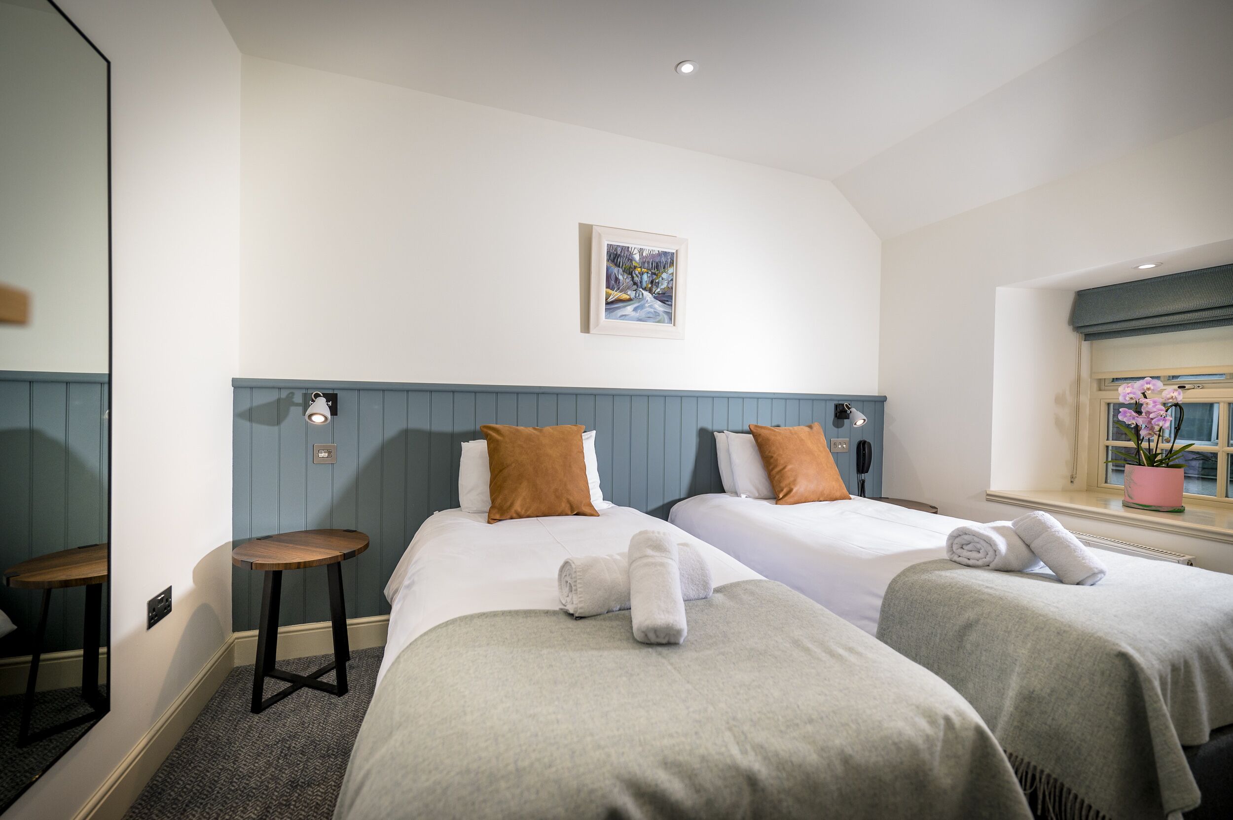 Une chambre d’hôtel avec deux lits, un petit tabouret, un tableau encadré au mur et une fenêtre avec une plante en pot.