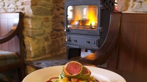A plate of food with a slice of meat and vegetables on a white plate, set on a wooden table with a stone wall and a wood-burning stove in the background.