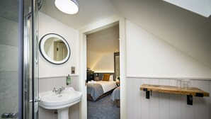 A bathroom with a sink, mirror, and shower. A bedroom is visible through an open door, featuring a bed and a bedside table.