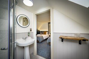 A bathroom with a sink, mirror, and shower. A bedroom is visible through an open door, featuring a bed and a bedside table.