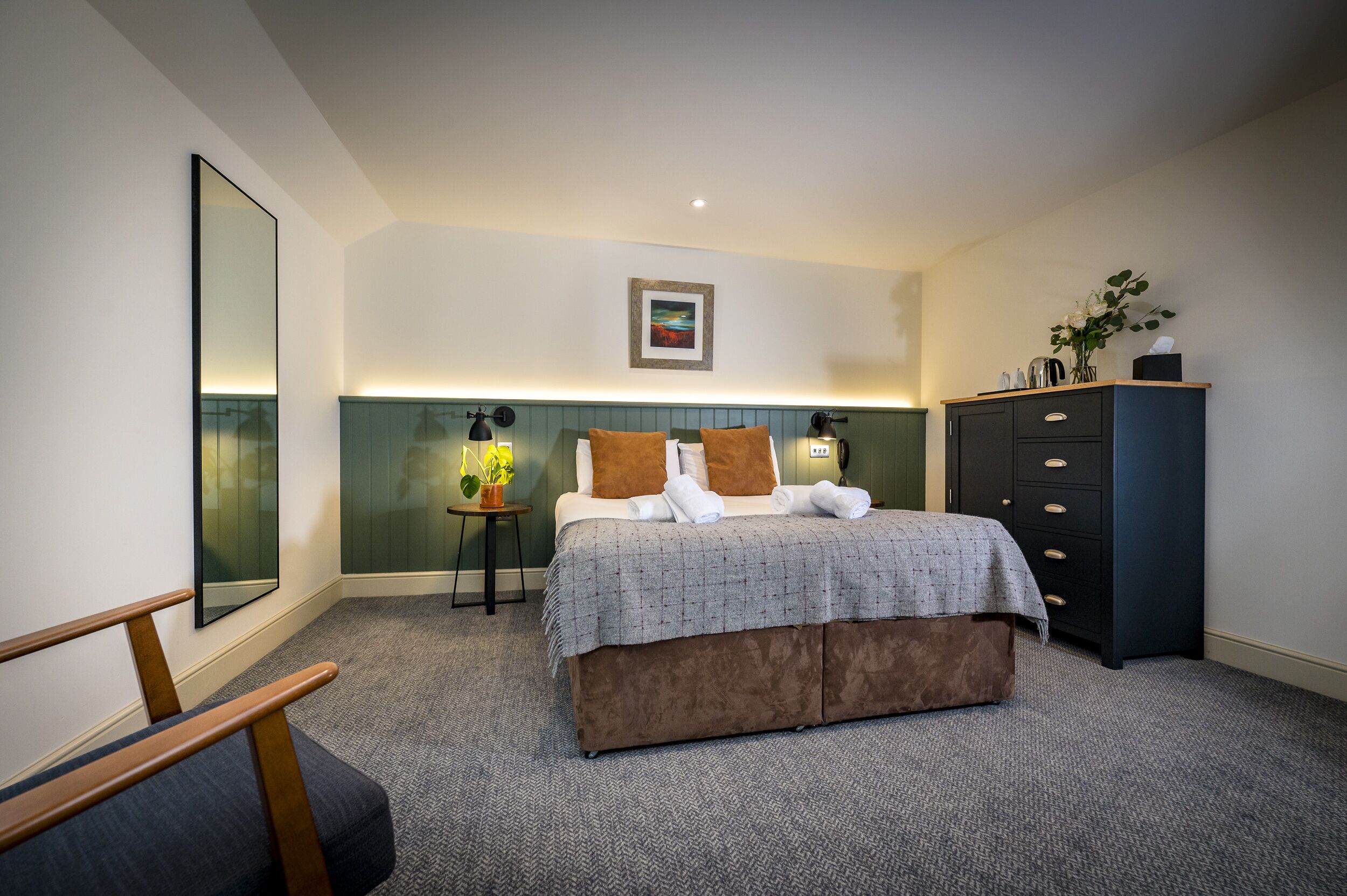 A hotel room with a bed, a dresser, a chair, a small table with a plant, and a framed picture on the wall.