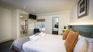 A hotel room with a large bed, a flat-screen TV mounted on the wall, a bathroom visible through an open door, and a window with blinds.