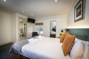 A hotel room with a large bed, a flat-screen TV mounted on the wall, a bathroom visible through an open door, and a window with blinds.