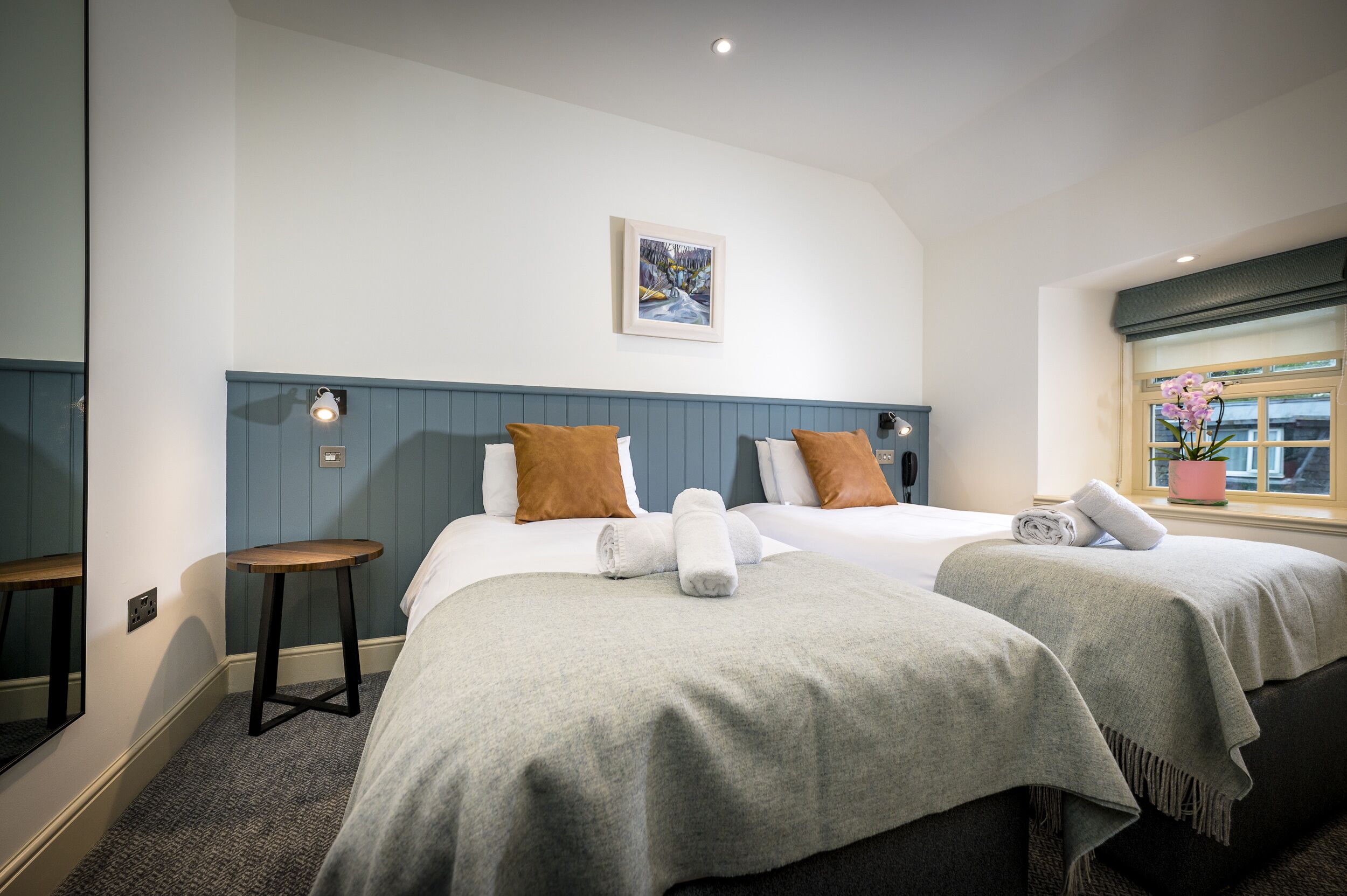 A hotel room with two beds, a small stool, a painting on the wall, and a window with a plant.