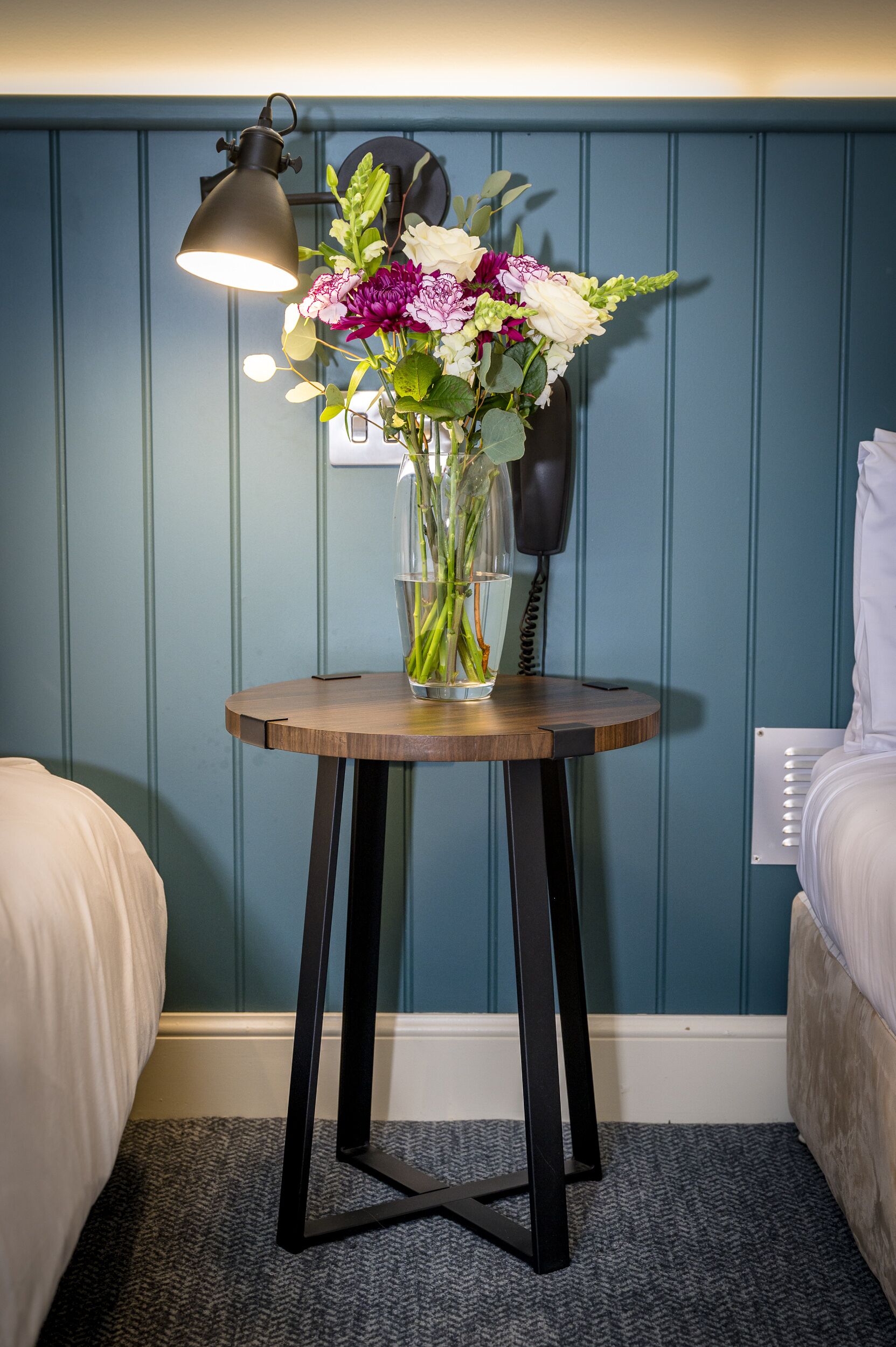 Una habitación con una mesa de madera que sostiene un florero con flores, una lámpara y un teléfono.