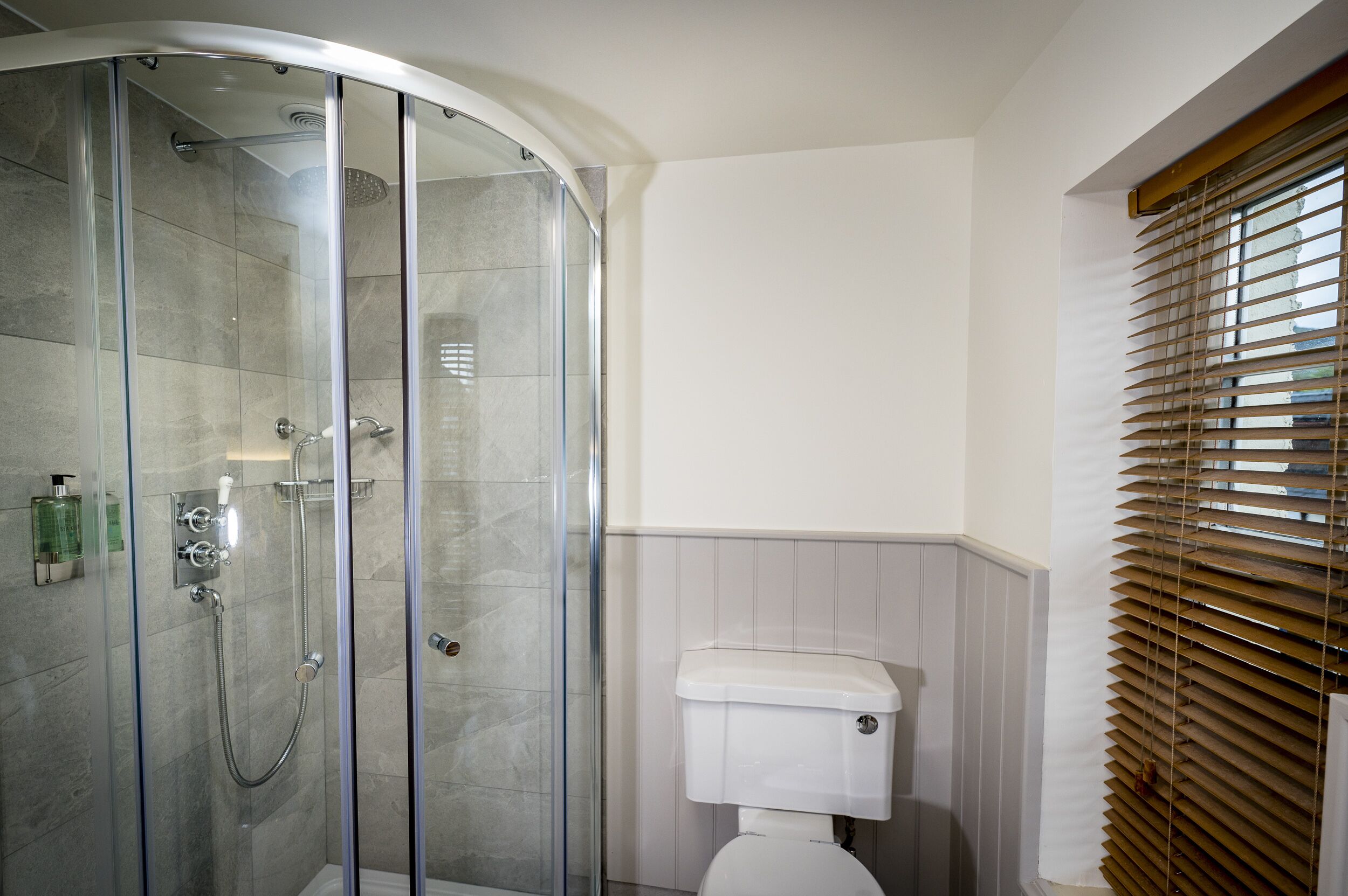 A bathroom with a glass-enclosed shower, a toilet, and a window with blinds.