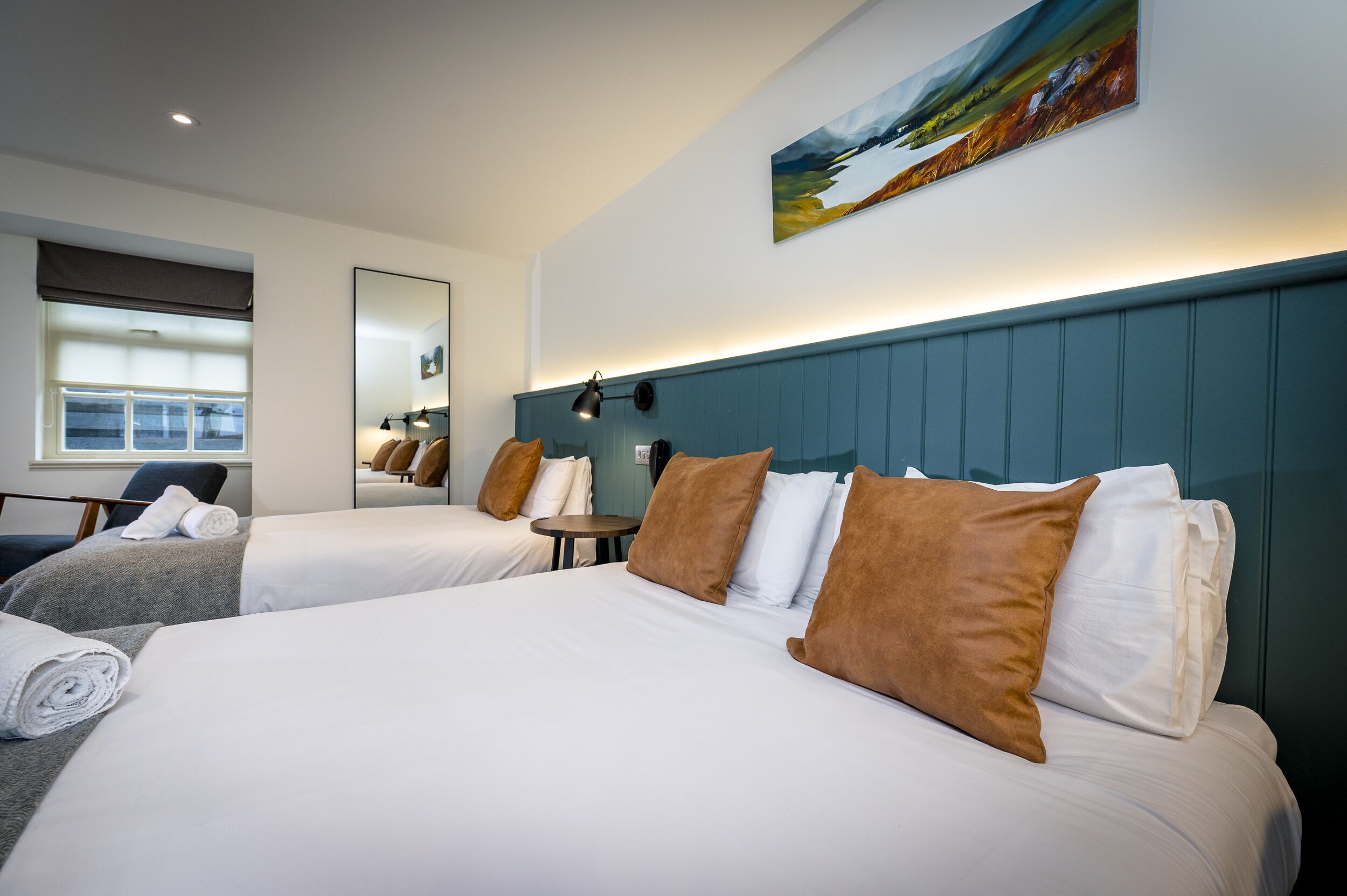 Habitación de hotel con dos camas, una mesita redonda pequeña, una silla, un espejo grande y un cuadro en la pared.