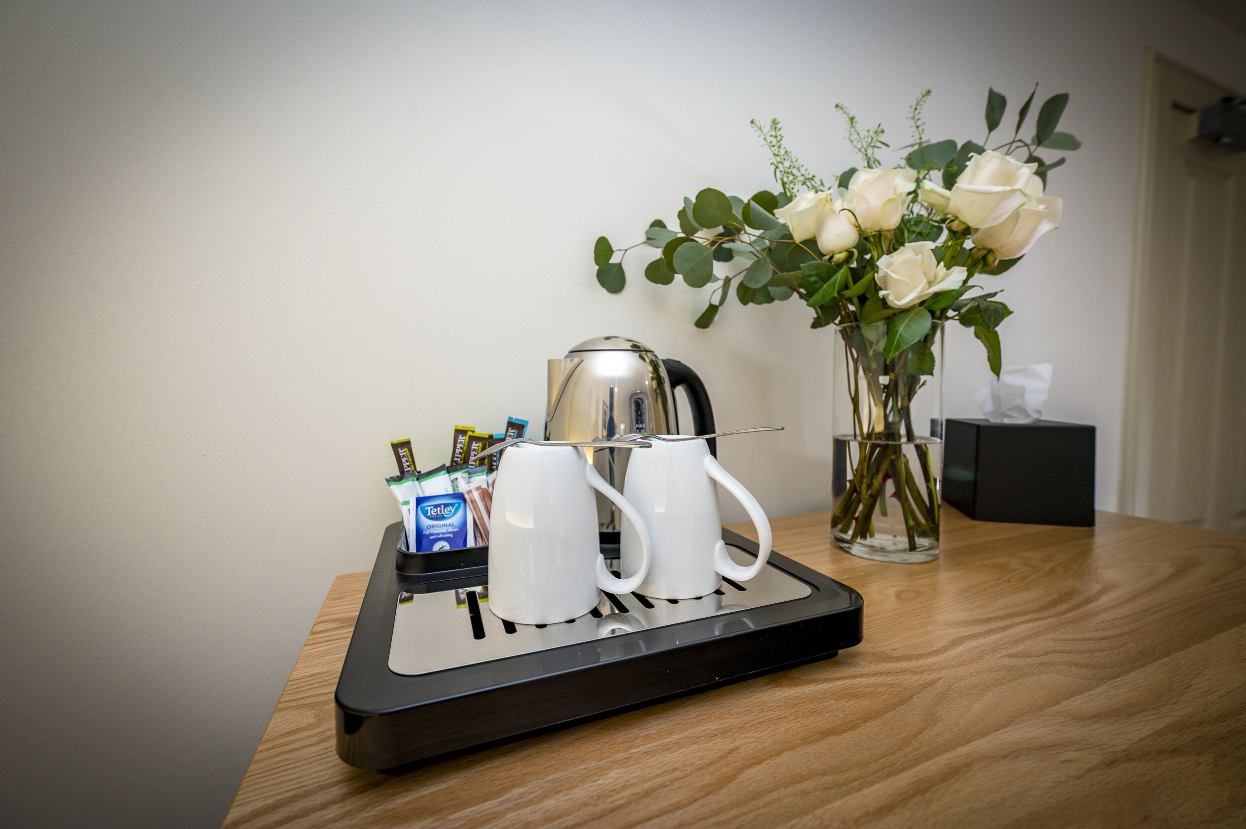 Een koffiehoek met een waterkoker, twee witte mokken en een vaas met witte rozen op een houten tafel.