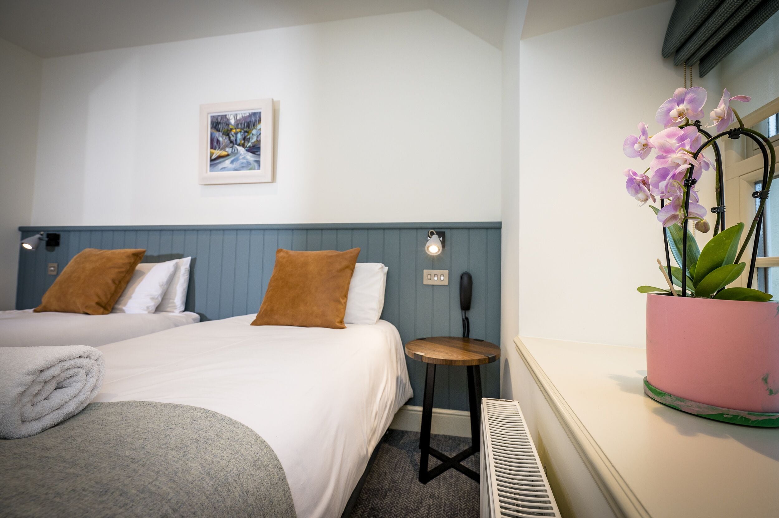 Quarto de hotel com duas camas, uma mesinha, um vaso de planta e um quadro na parede.