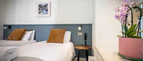 A hotel room with two beds, a small table, a potted plant, and a painting on the wall.