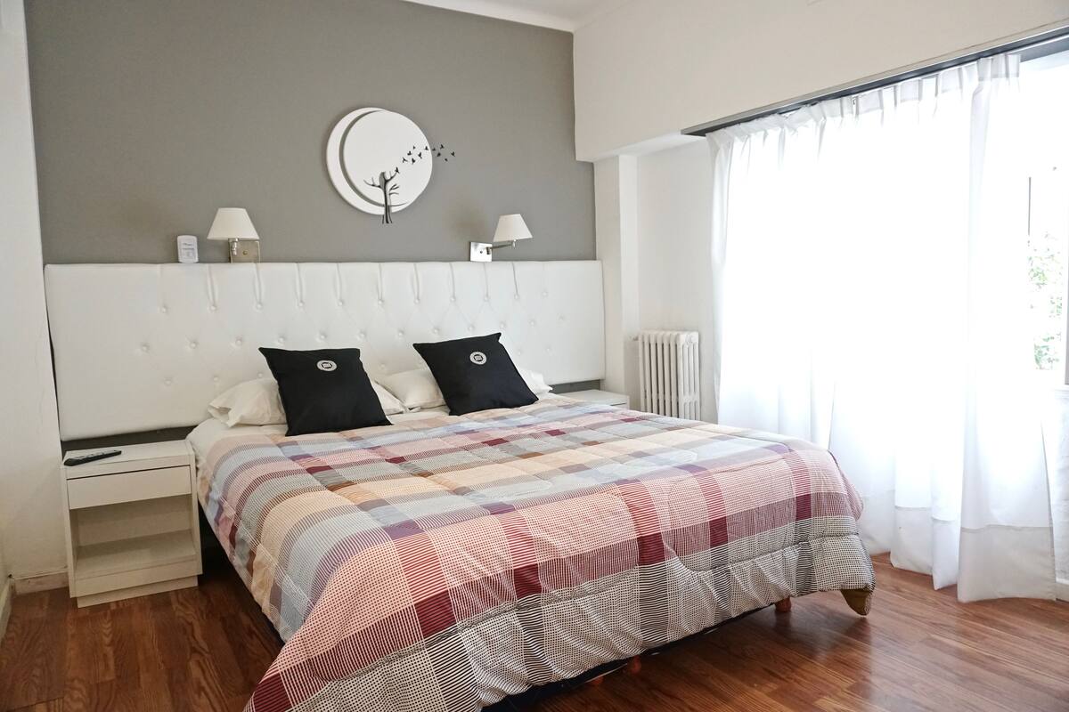 A bedroom with a large bed, two bedside tables, a wall clock, and a window with curtains.