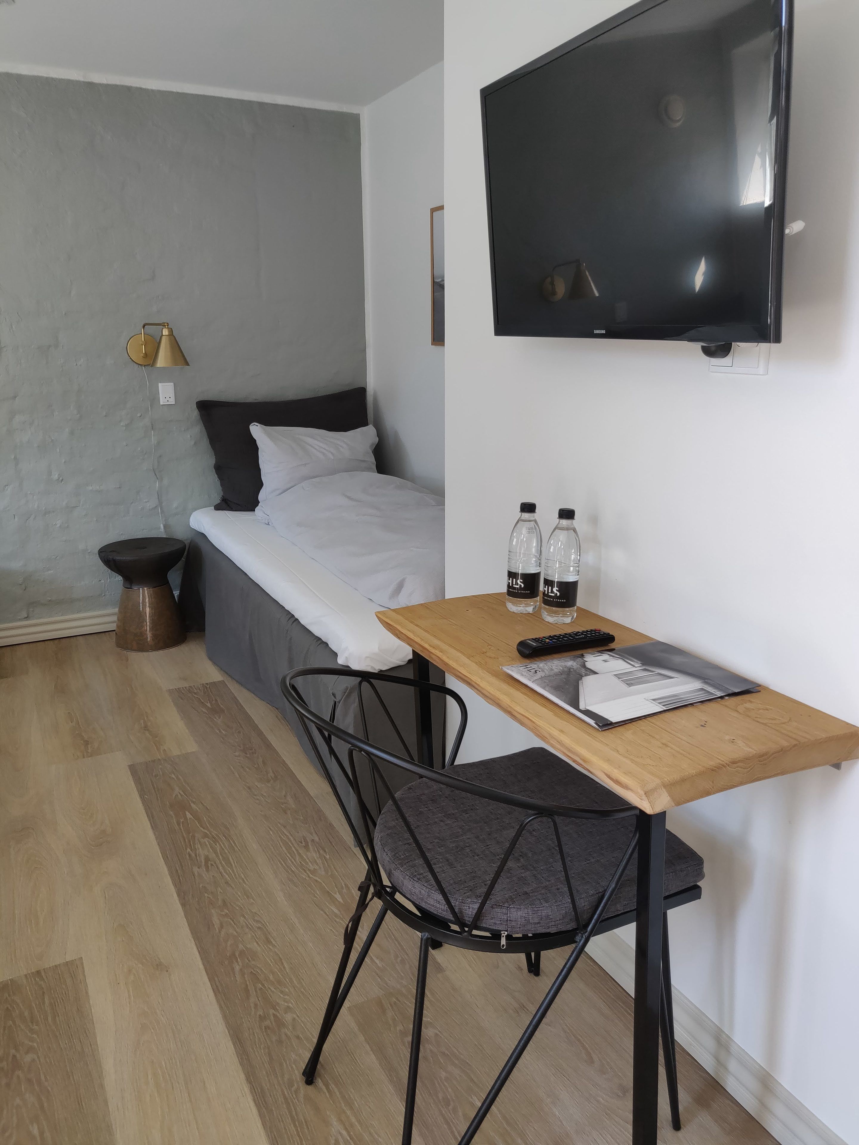 Een kleine, moderne kamer met een bed, een aan de muur bevestigde televisie, een houten tafeltje met twee flessen en een afstandsbediening, en een stoel.