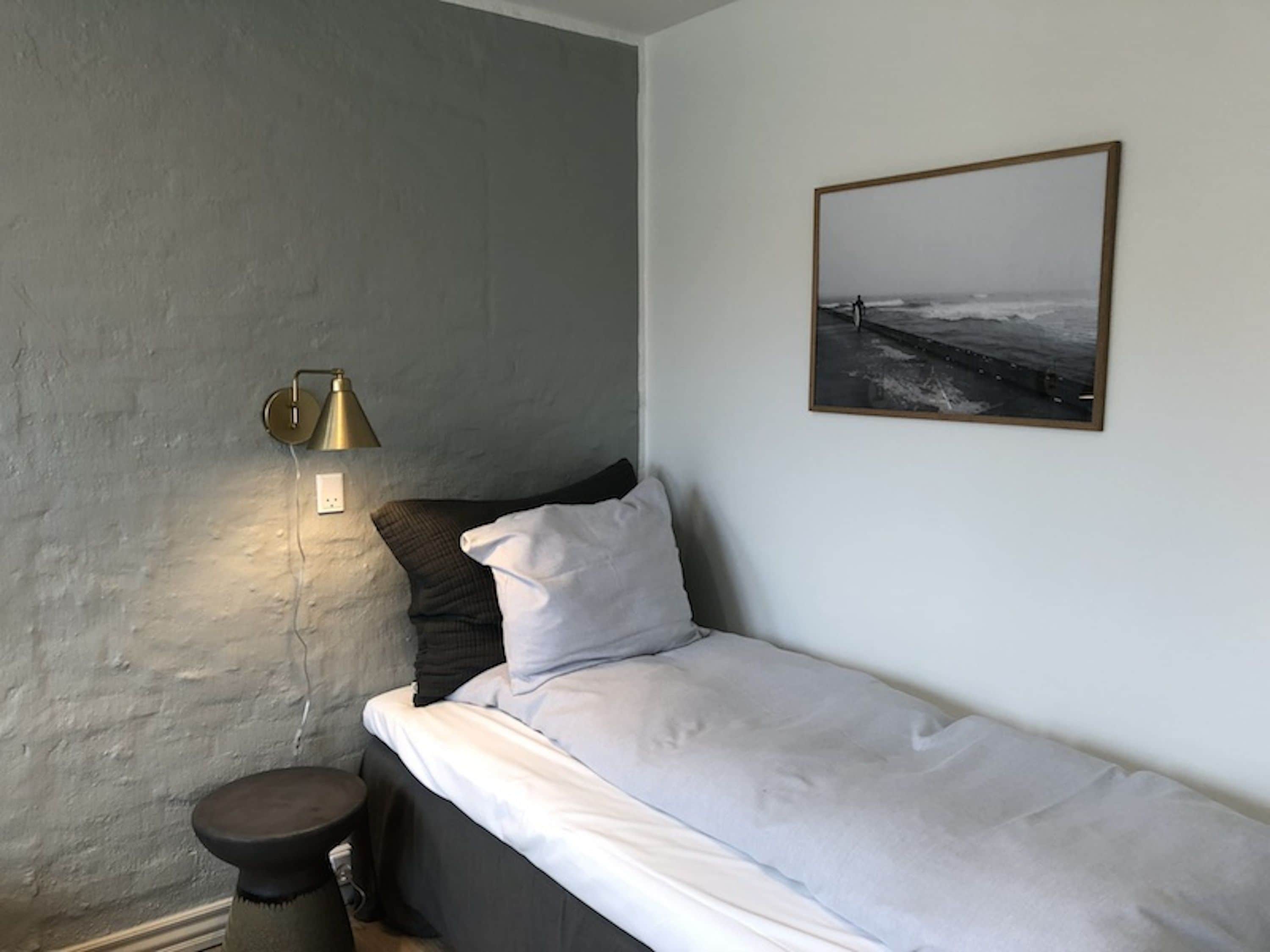 En enkeltseng med hvide sengetøj, en mørk pude og en lille skammel. En vægmonteret lampe er synlig på venstre side.