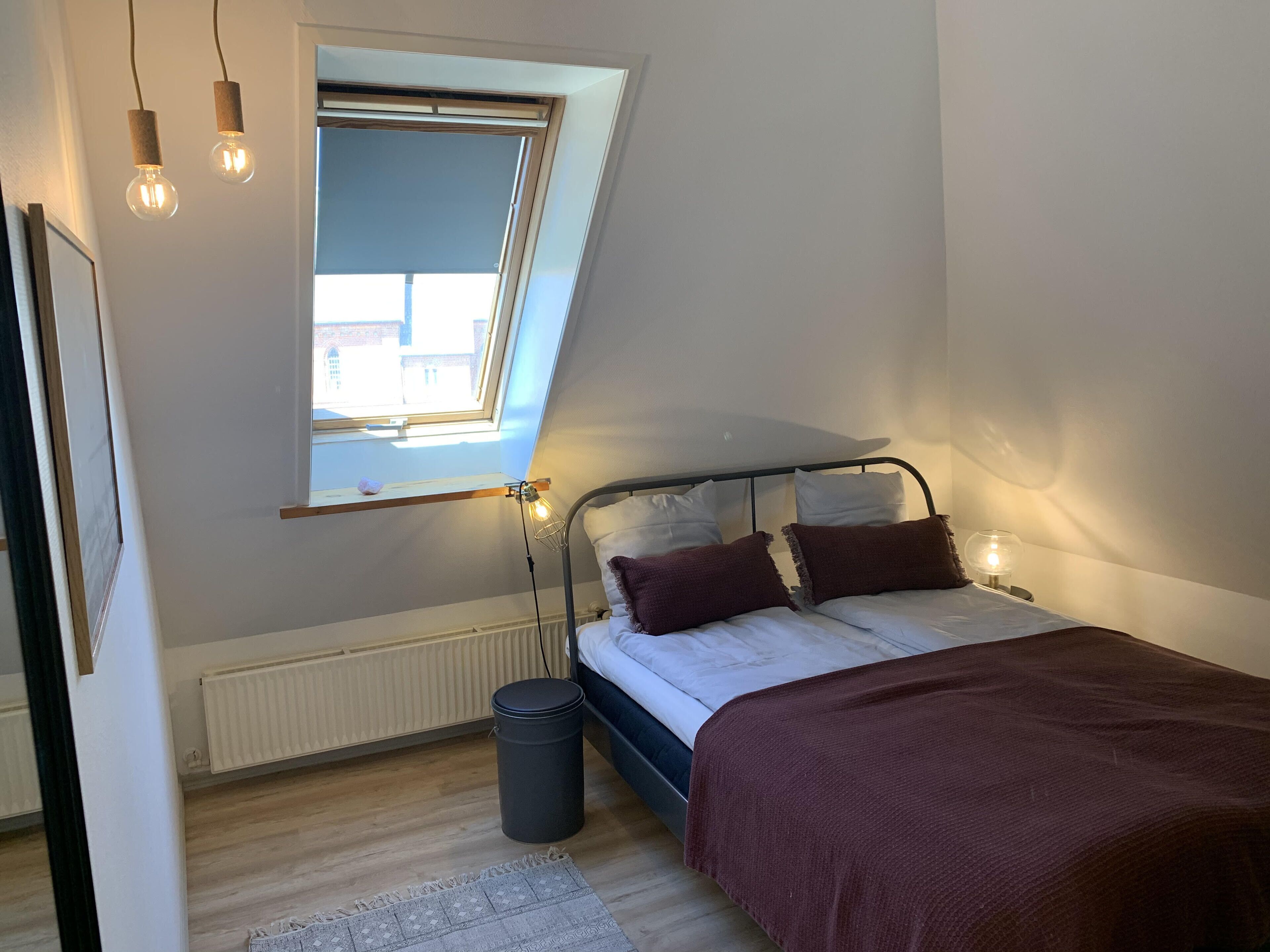 A bedroom with a sloped ceiling, a skylight, a bed with a headboard, bedside lamps, a radiator, and a trash bin.