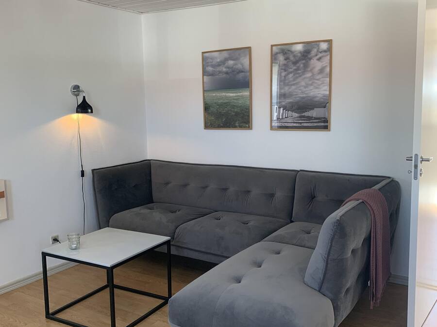 A modern living room with a grey sectional sofa, a small white table, and a floor lamp. There are two framed pictures on the wall.