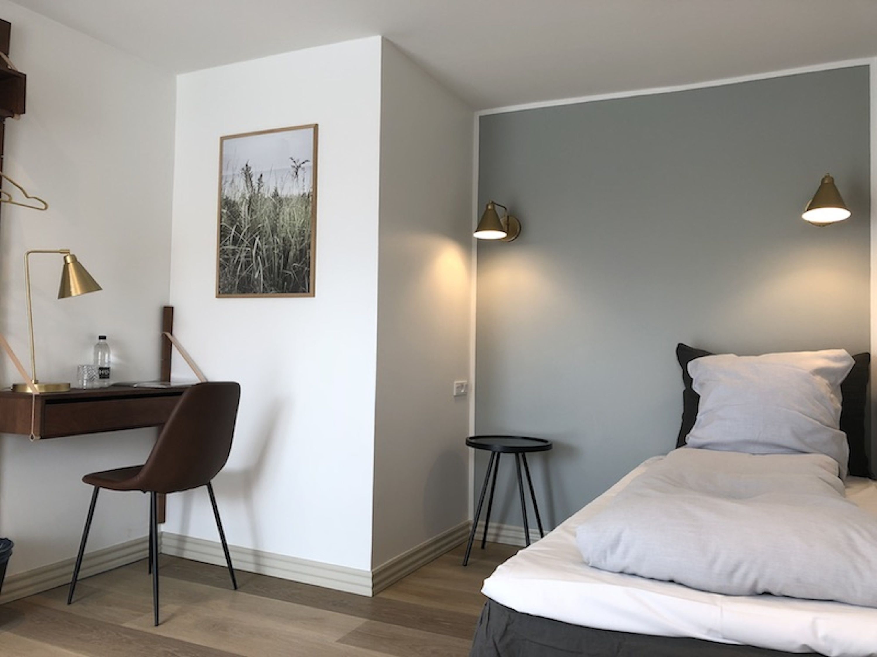A hotel room with a bed, a desk, a chair, a small stool, and a wall-mounted lamp.