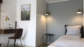 A hotel room with a bed, a desk, a chair, a small stool, and a wall-mounted lamp.