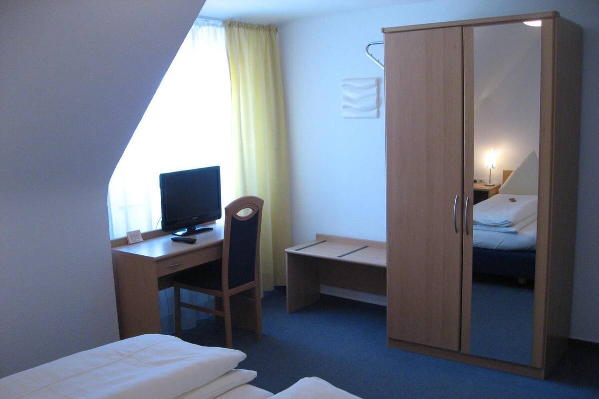 A hotel room with a single bed, a desk with a computer, a chair, a wardrobe, and a small bedside table with a lamp.